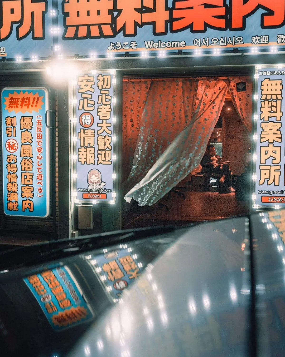 Nighttime view of an entrance to a Japanese establishment with illuminated signage in Japanese, Korean, and English, displaying welcoming messages. The entrance has a floral curtain, and inside, a person is sitting on a bench with another person near