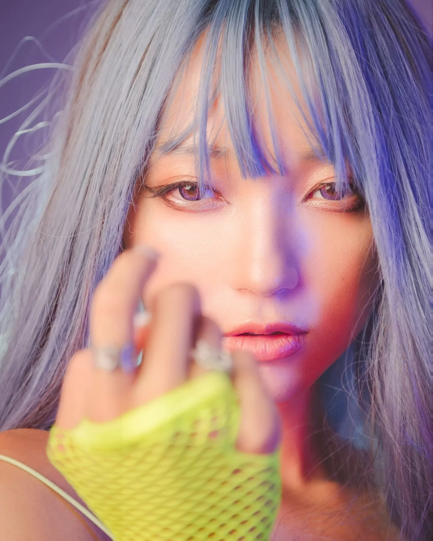 Close-up of a woman with silver and purple hair, eye makeup, and a neon green fishnet glove, holding her hand near her face.