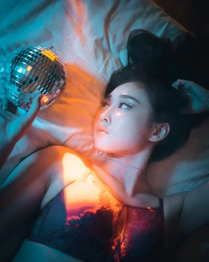 A woman lying on her back on a bed, looking at a small disco ball held by a hand, with colorful, reflective lighting illuminating her face and surroundings.