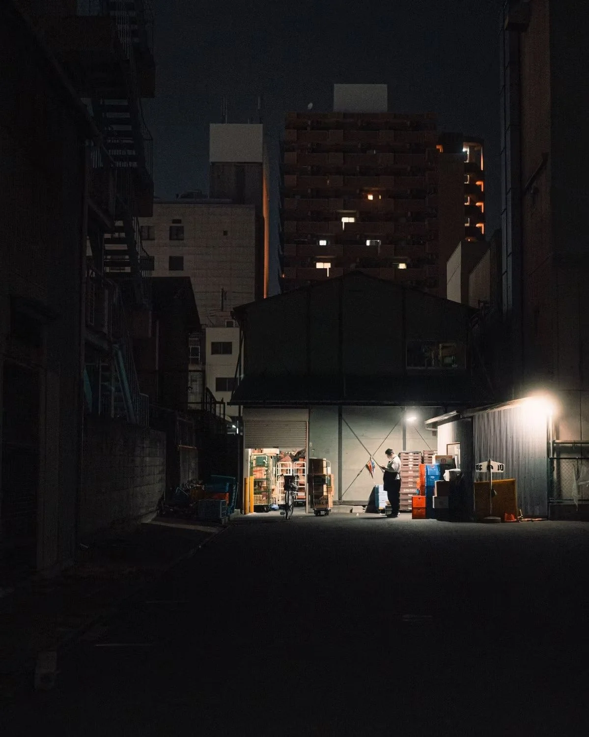 Nighttime scene of a person standing outside a warehouse, illuminated by artificial lights, with storage shelves, carts, and boxes in the background, surrounded by tall buildings.