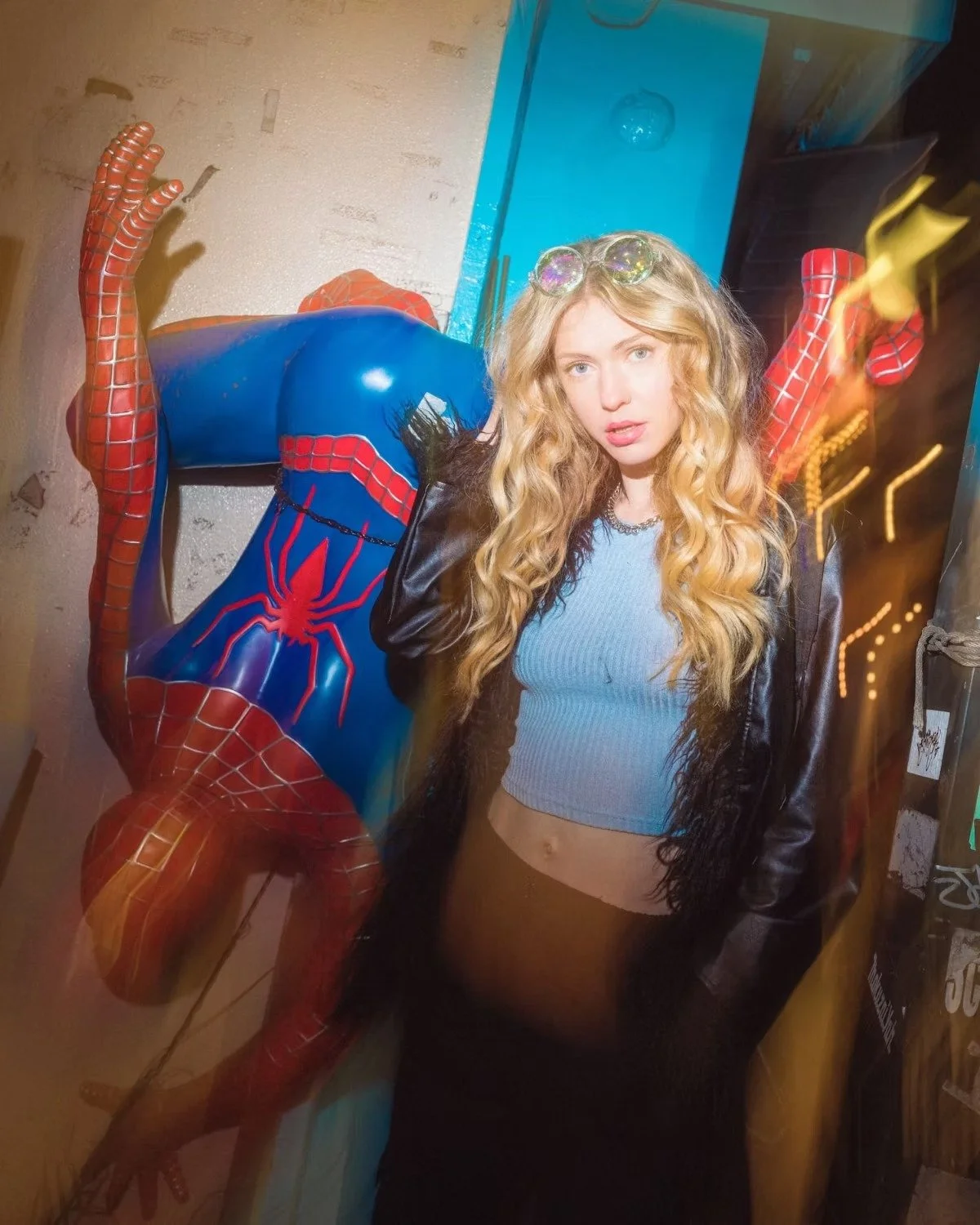 A young woman with long blonde hair poses next to a Spider-Man statue in a vibrant urban setting. The motion blur and colorful lights enhance the playful and dynamic street atmosphere. Street photography by Deniz Demir.
