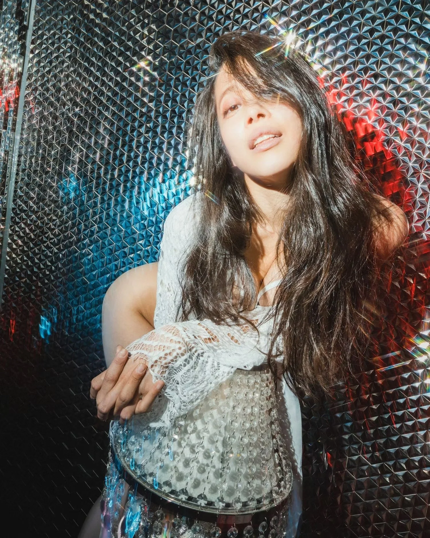 A woman with long dark hair is posing in front of a shiny, geometric, reflective backdrop, wearing a white lacy top. Bright lights create starburst effects and rainbow reflections around her.