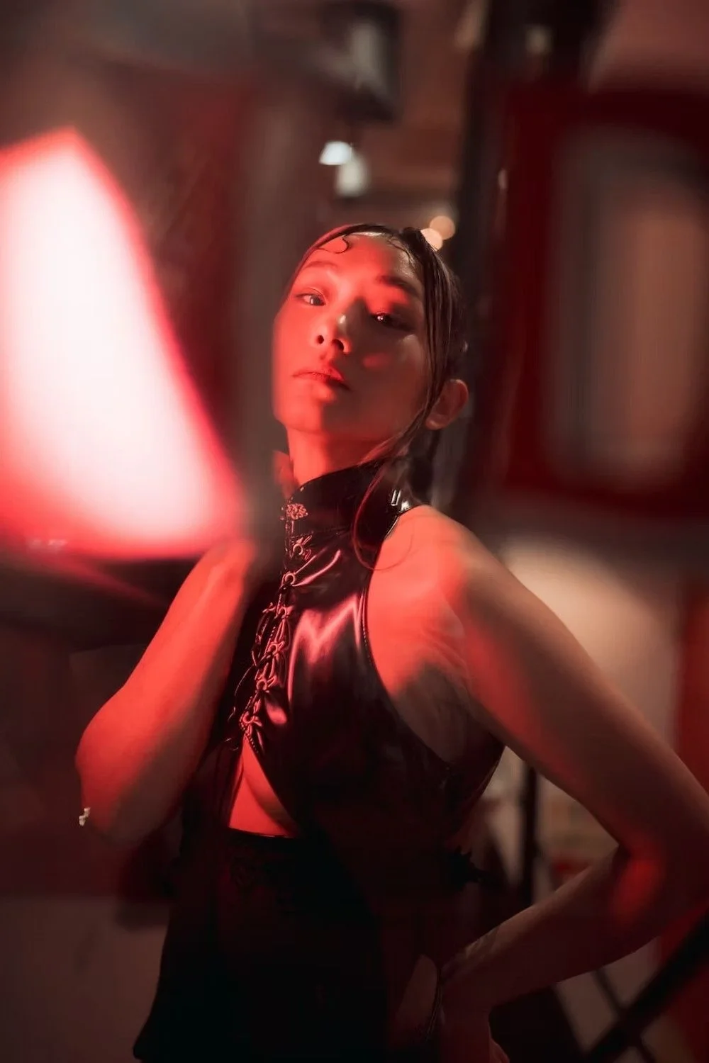A woman with dark hair styled in waves, wearing a shiny black leather dress, posing with a confident expression in a dimly lit environment with red lighting in a Tokyo alley.