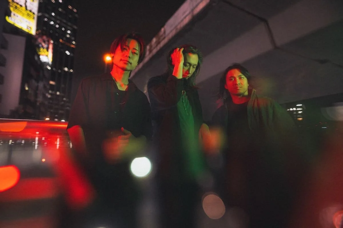 Three young men standing under a city bridge at night with colorful lights reflecting on their faces.