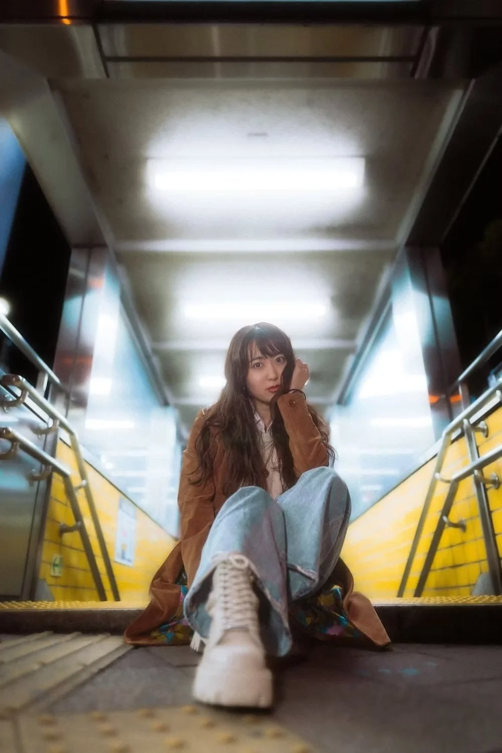 A young woman with long wavy hair, wearing a brown coat, white shirt, and jeans, sitting on the floor of an elevator with yellow walls and metal handrails, looking at the camera.