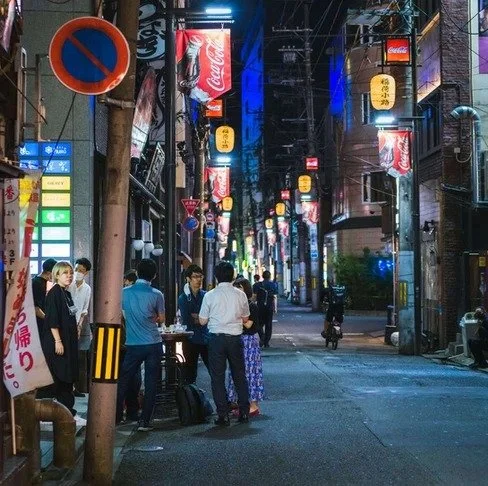 A group of people gathers under glowing lanterns in a narrow Tokyo alley, as neon signs and streetlights reflect off the wet pavement at night.
