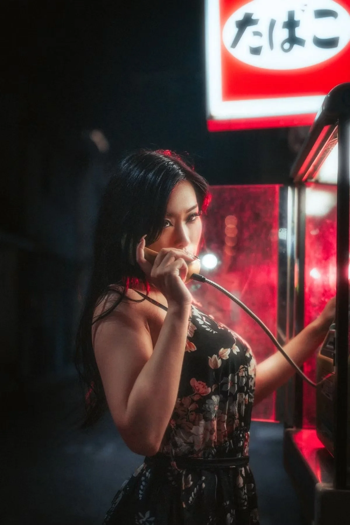 A woman with dark hair in a floral dress is using a payphone at night, with neon signs in the background shot in Osaka Japan.