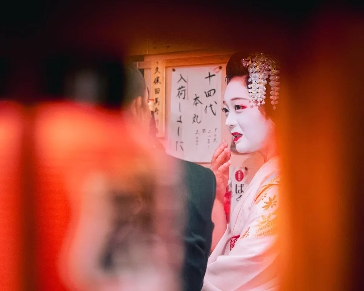 A young woman dressed as a traditional Japanese geisha, wearing white face makeup, red lipstick, and an ornate hairpiece with flowers and beads, engaging in conversation in a setting with Japanese calligraphy on a wooden board in the background. Shot