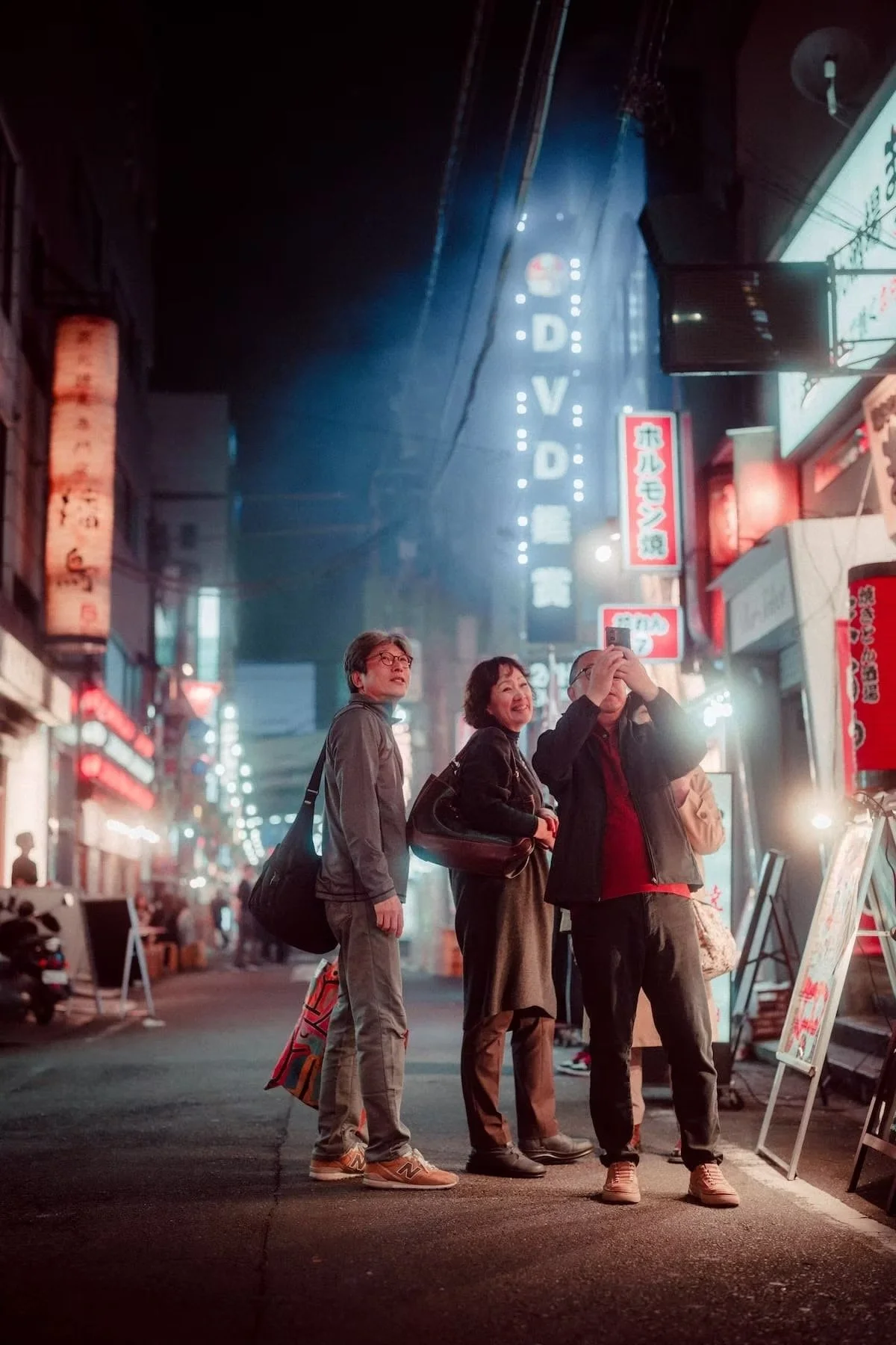 Three Japanese people standing on a busy city street at night, with illuminated signs in Japanese. One is taking photos or videos with a phone, while the others look on on the streets of Shimbashi in a cyberpunk scene.