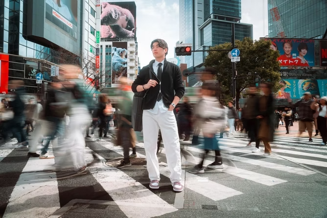 Young man standing in the middle of a busy city crosswalk wearing a black blazer, white pants, and holding a phone, surrounded by blurred pedestrians and large digital billboards.