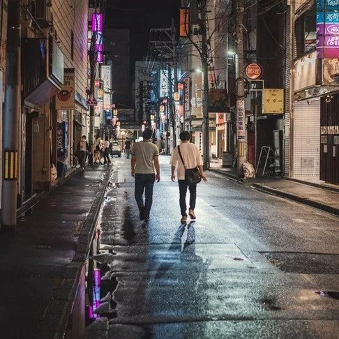 A couple walks down a wet Tokyo street at night, neon lights reflecting on the pavement, creating a moody and cinematic urban atmosphere.
