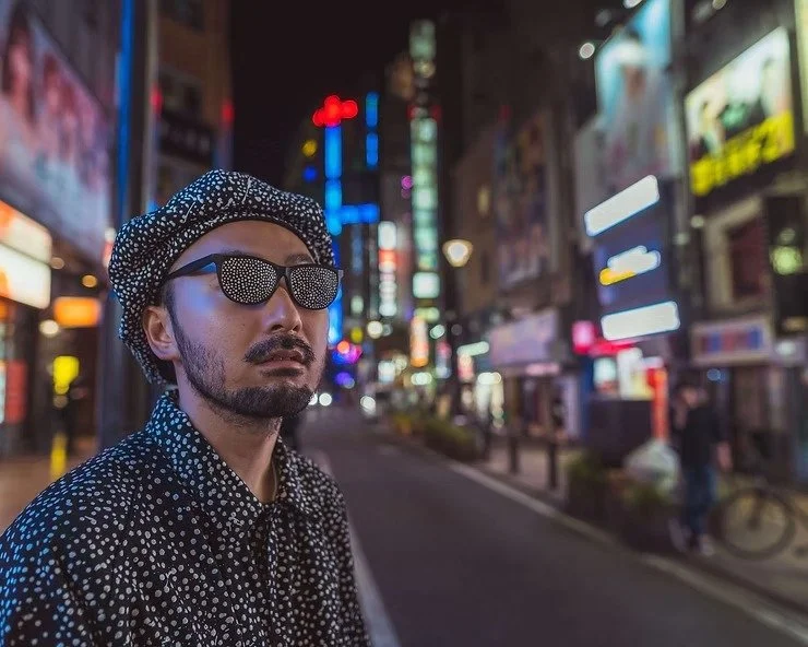 A man in a polka-dot outfit and sunglasses, standing on a lit street in Kabukicho, with neon signs blurred in the background.