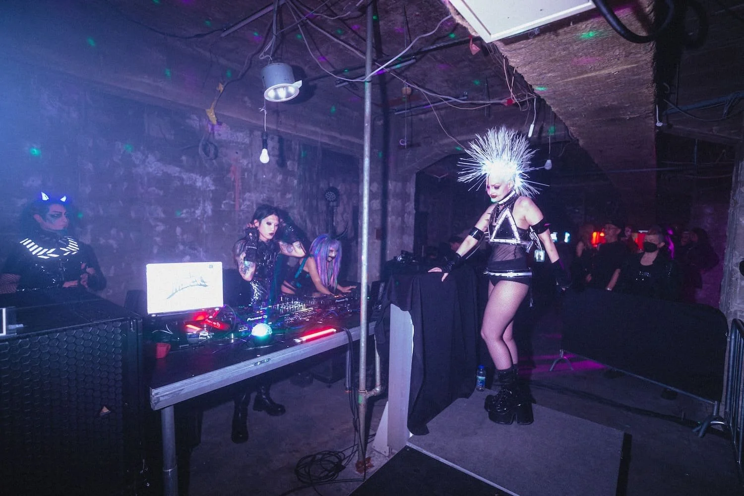 Bondage Ball 
---
Performance at a nightclub with a DJ and a performer dressed in goth and punk-inspired attire, including a spiky white wig, fishnet stockings, and platform boots, in a dimly lit setting with stone walls and colorful lights.