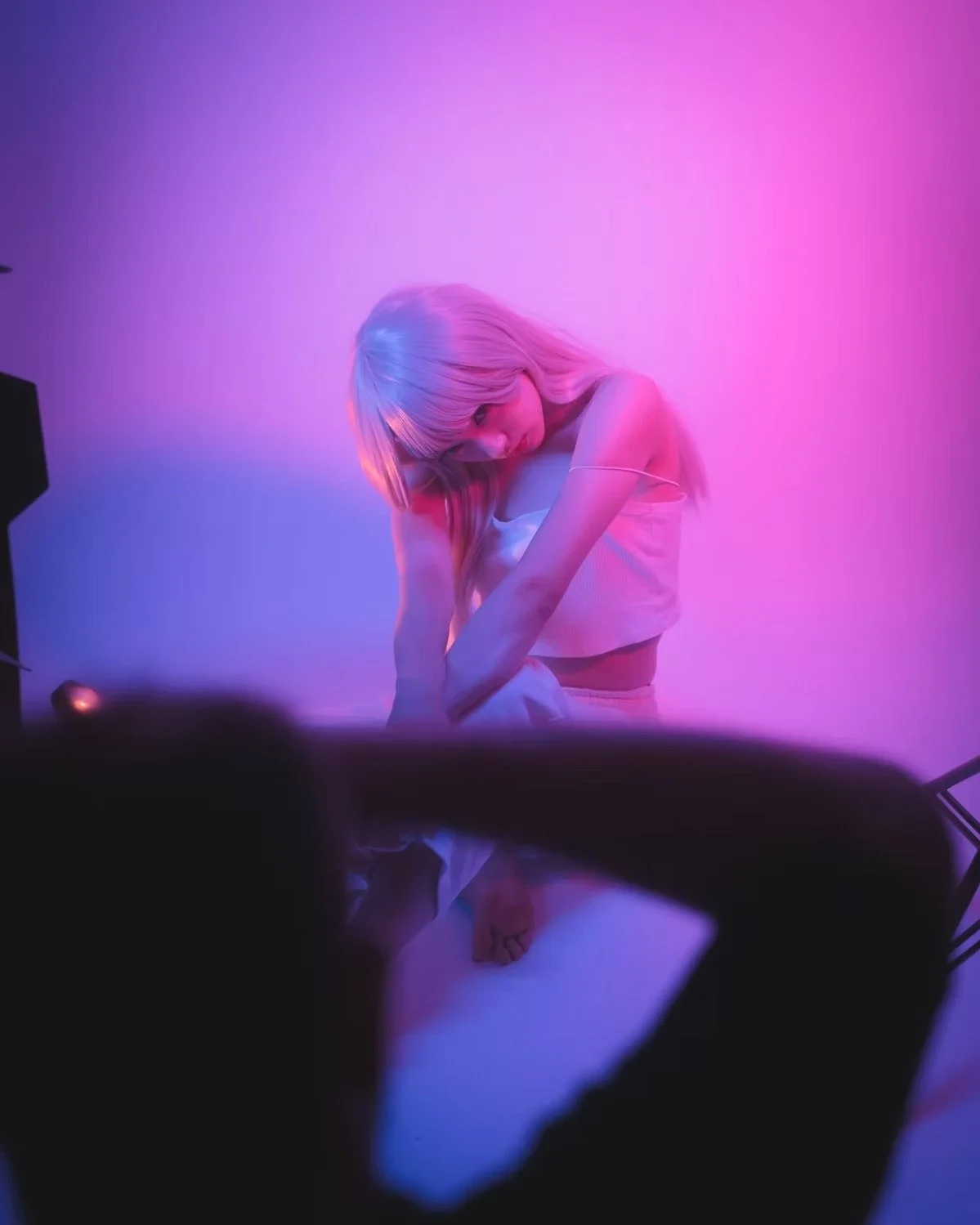 A woman with blonde hair in a white top is posing against a pink and blue gradient background, illuminated by colored lights.
