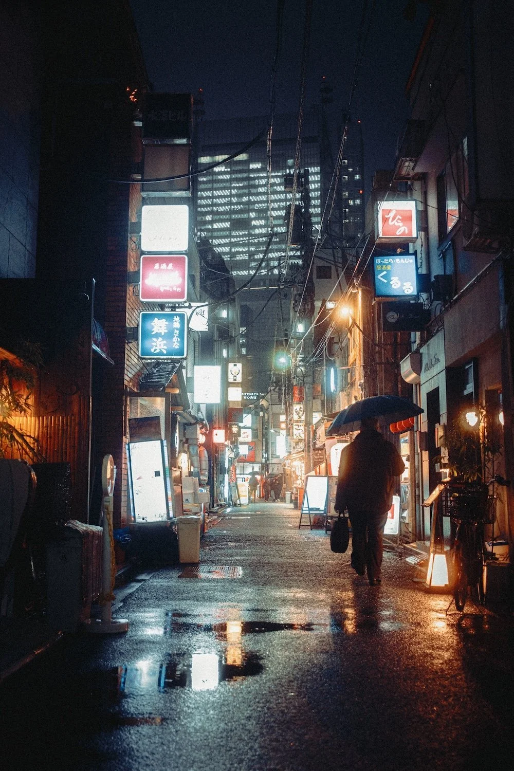 A lone salaryman holding an umbrella walking through a wet street at night in Shimbashi, Tokyo, illuminated by colorful neon signs in Japanese, with buildings and power lines overhead.