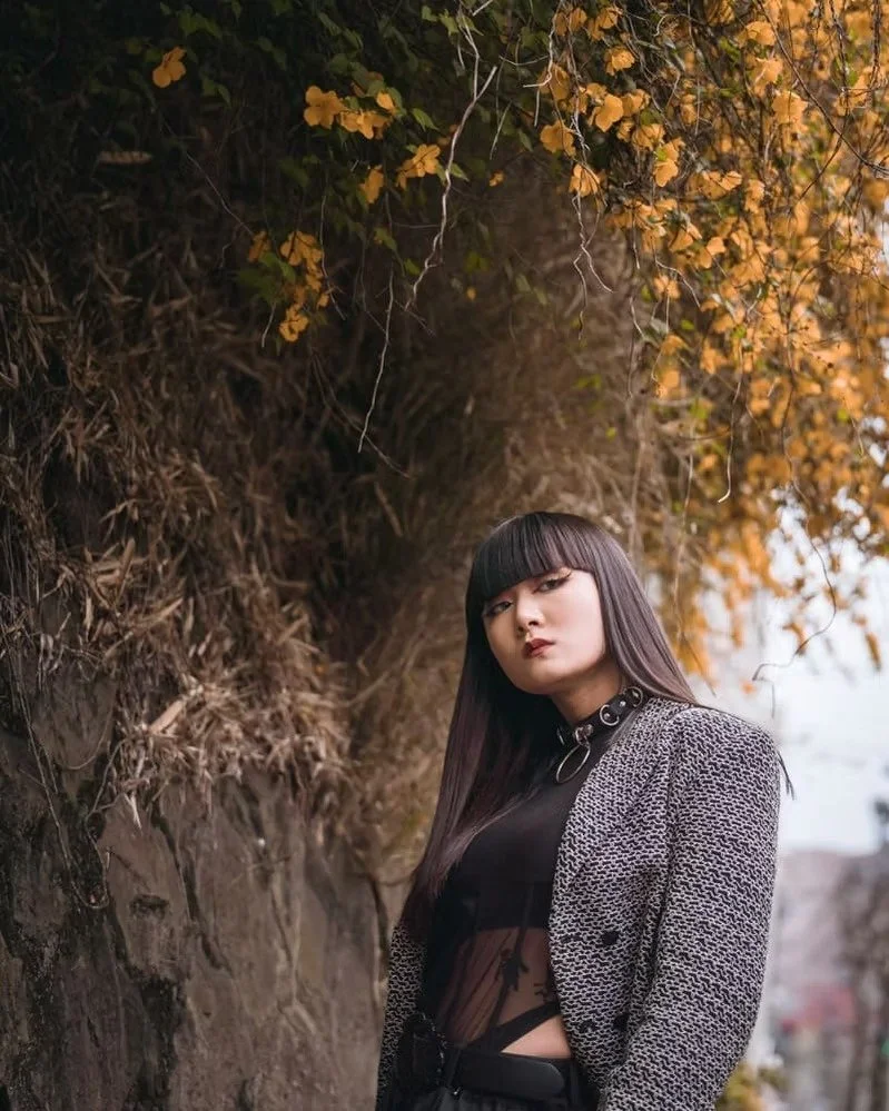 A woman with long dark hair and bangs posing outdoors against a stone wall and autumn foliage, wearing a black top with a sheer middle section and a patterned blazer.