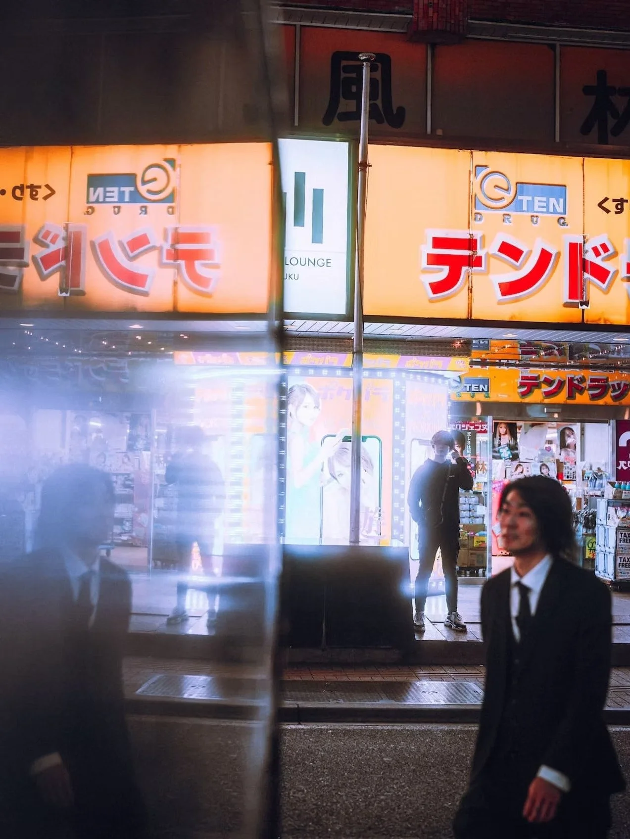 Nighttime street scene in Japan with illuminated signs, people walking by, some wearing masks and formal attire, storefronts with advertisements and posters, and a window reflecting images.