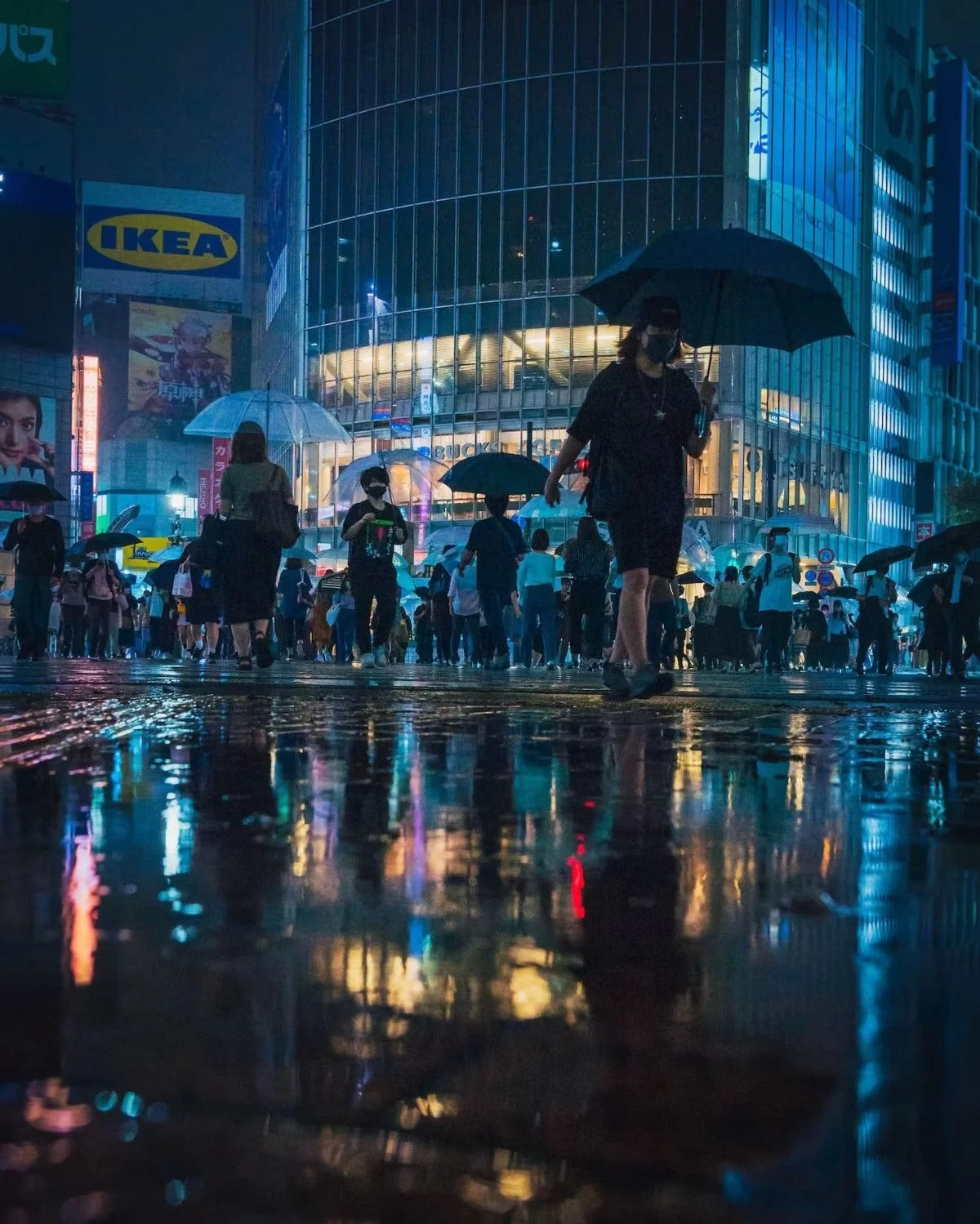 People with umbrellas walking in the rain at night in a city with illuminated buildings and reflections on wet pavement during the covid lockdown in Shibuya Tokyo.