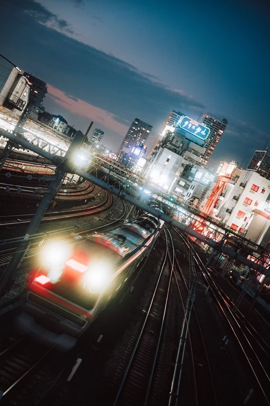 A Tokyo cityscape at night with illuminated buildings, railway tracks, and a train passing through with bright lights, under a dark sky.