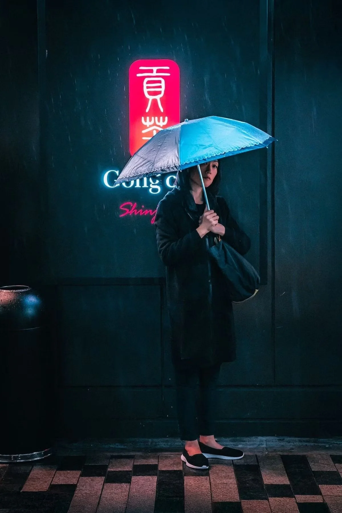 A woman holding a blue umbrella stands in front of a dark wall with neon signs, wearing a black coat and black shoes with white soles, during rainy weather in Shinjuku.