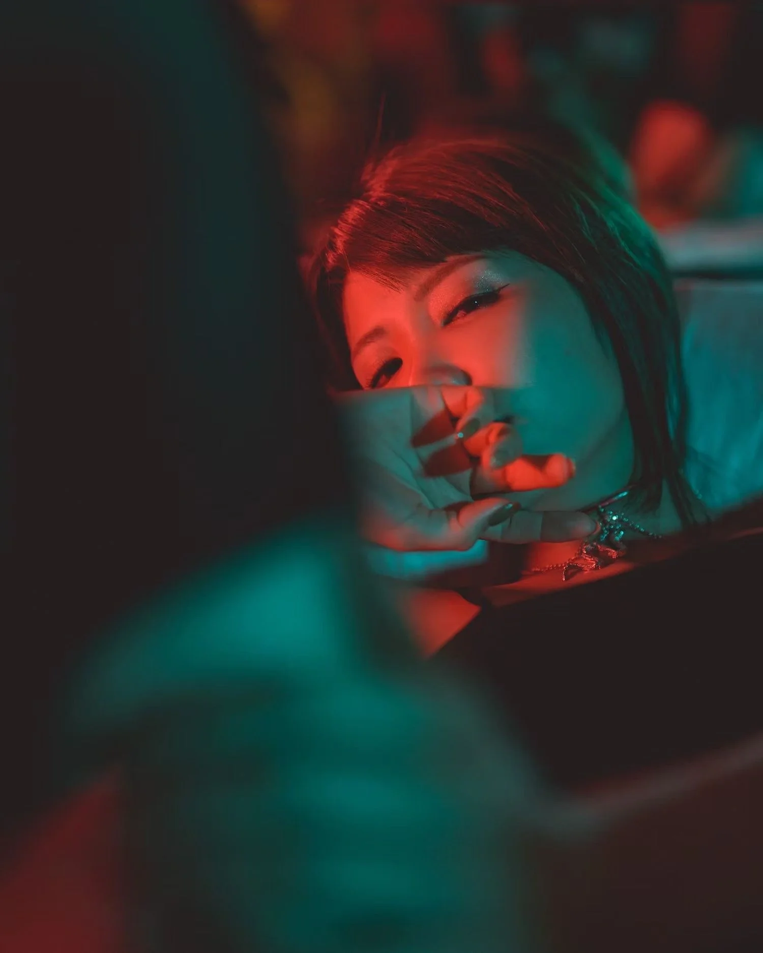 A woman with short hair lying down with her eyes closed, illuminated by red and green neon lights, with her hand partially covering her face.