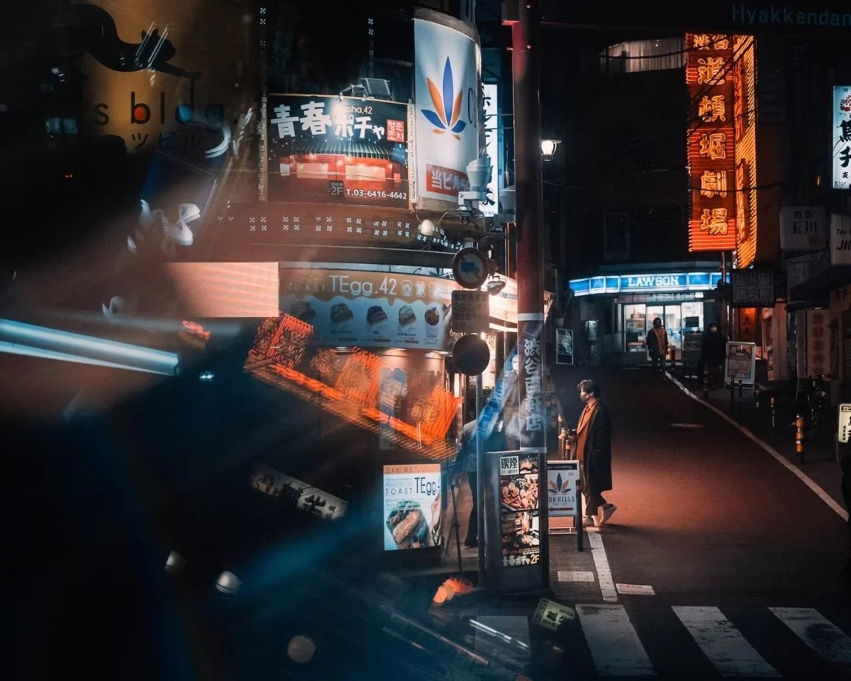 Night scene of a city street with bright neon signs and advertisements in Japanese and English. A person is standing at the sidewalk near the crosswalk, illuminated by the streetlights and signage. Street photography in Shibuya.