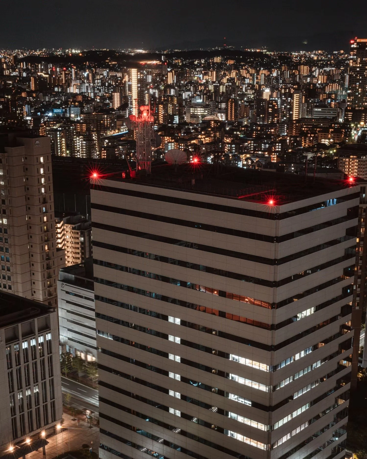 Nighttime cityscape with brightly lit skyscrapers and buildings, including a prominent high-rise with striped black and white exterior, with red lights on top indicating aircraft safety, and illuminated city streets below.