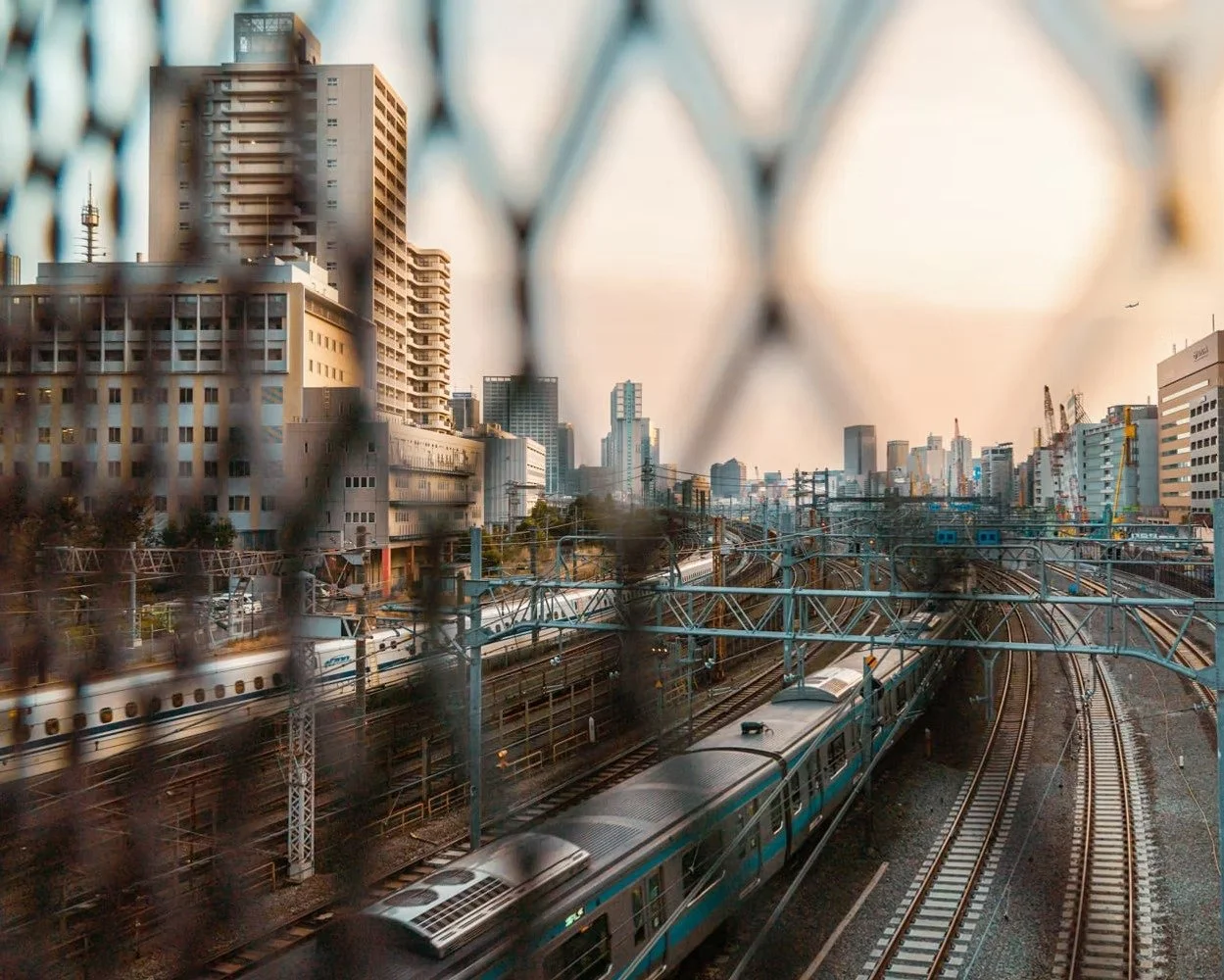 Cityscape with buildings, railway tracks, and a train seen through a chain-link fence.