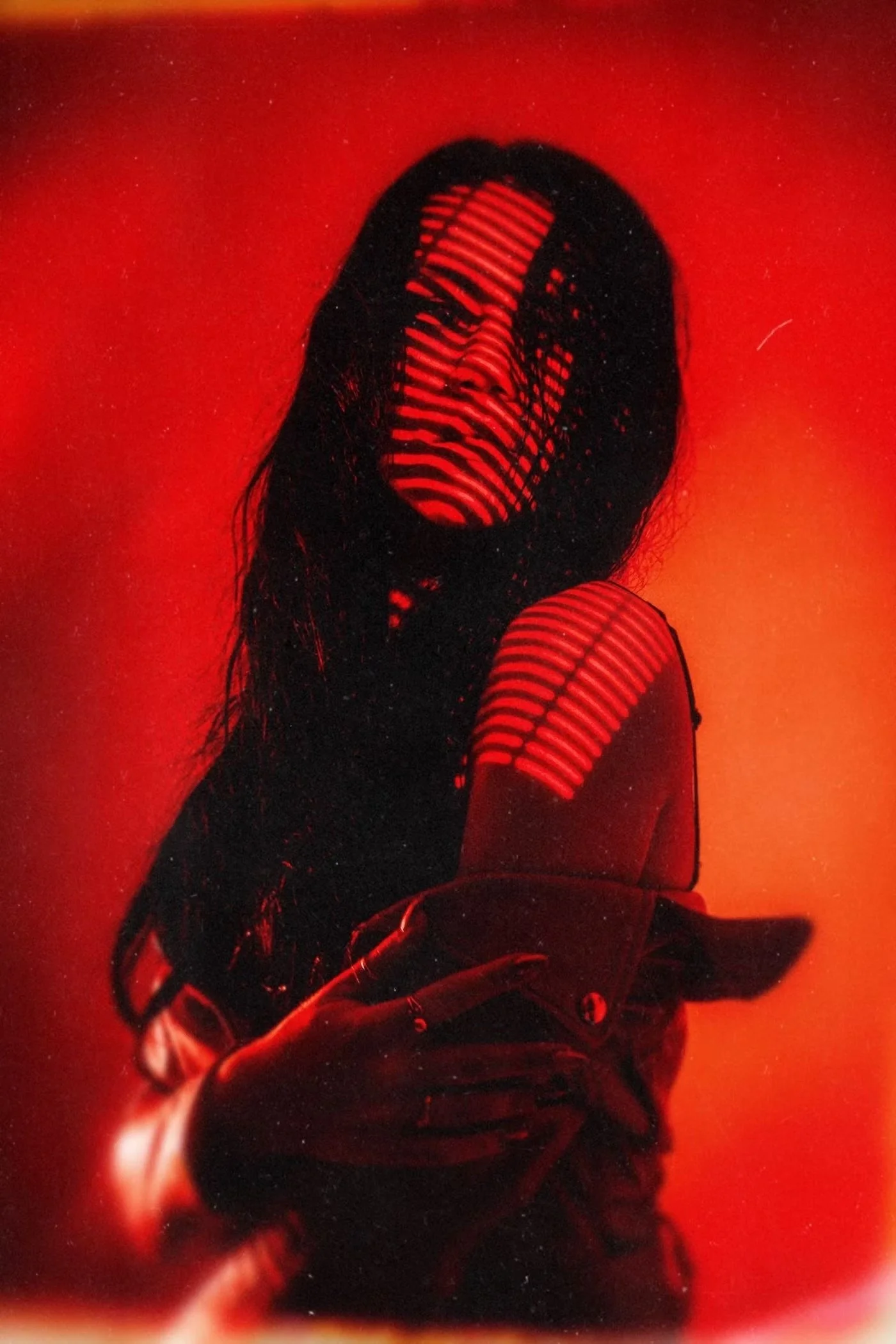 A person with long hair taking a selfie against a red backdrop, wearing a sleeveless striped top and shorts shot in Studio in Tokyo by Deniz Demir.