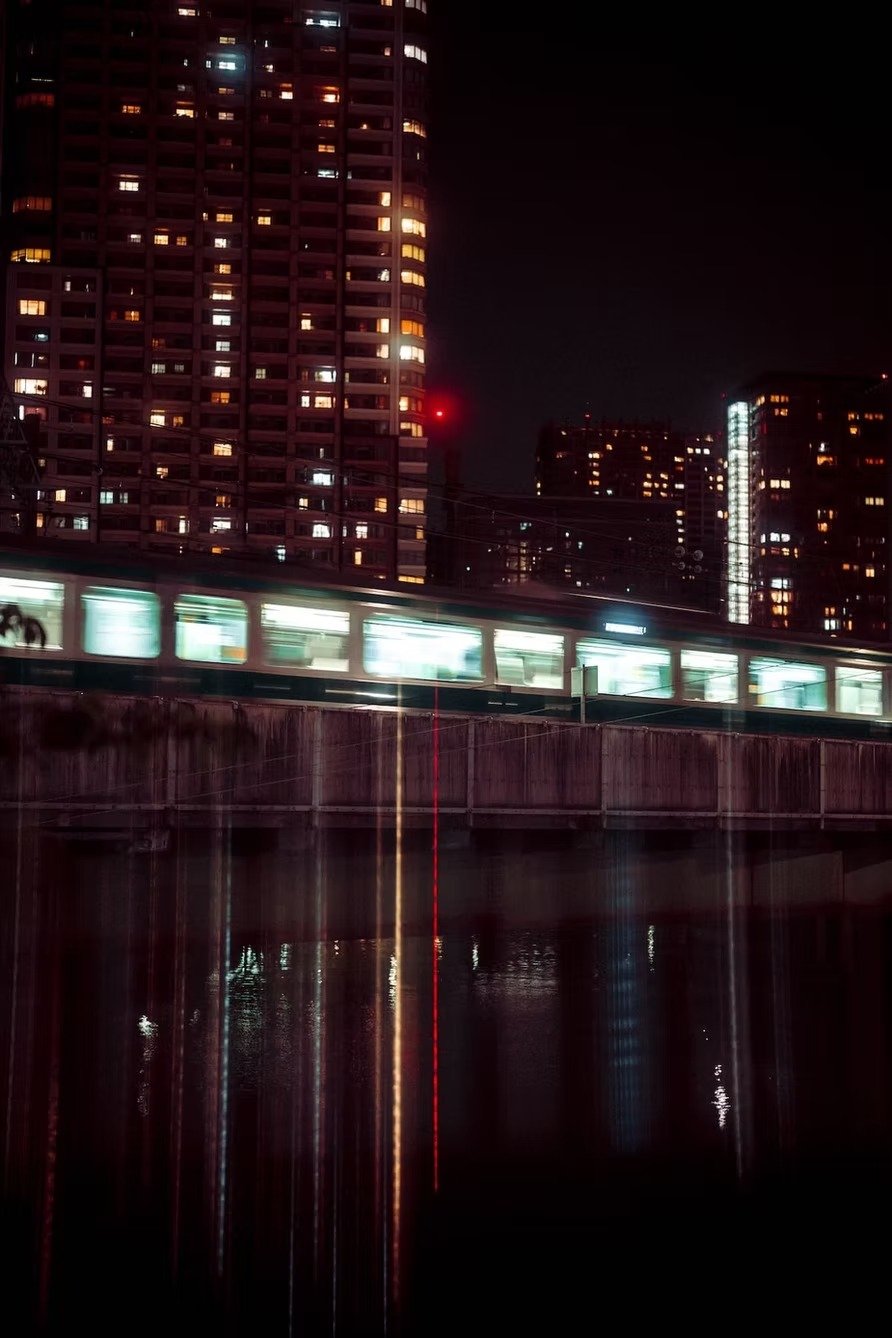 Night cityscape with illuminated high-rise buildings, a moving train on a bridge, and reflections in the water below.