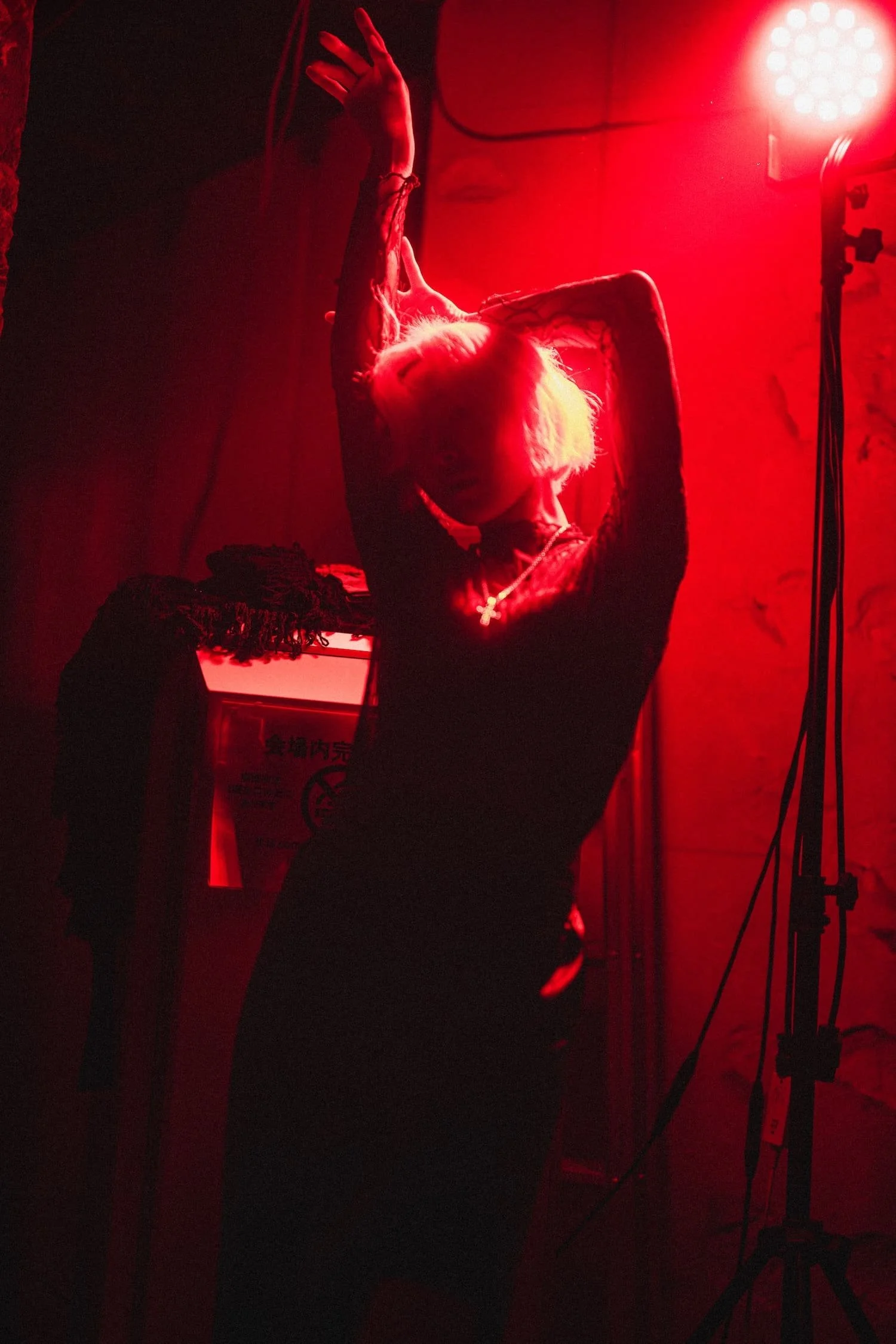Bondage Ball 
---
A person with platinum blonde hair and dark clothing posing with their hands above their head in a red-lit room, surrounded by professional lighting equipment.