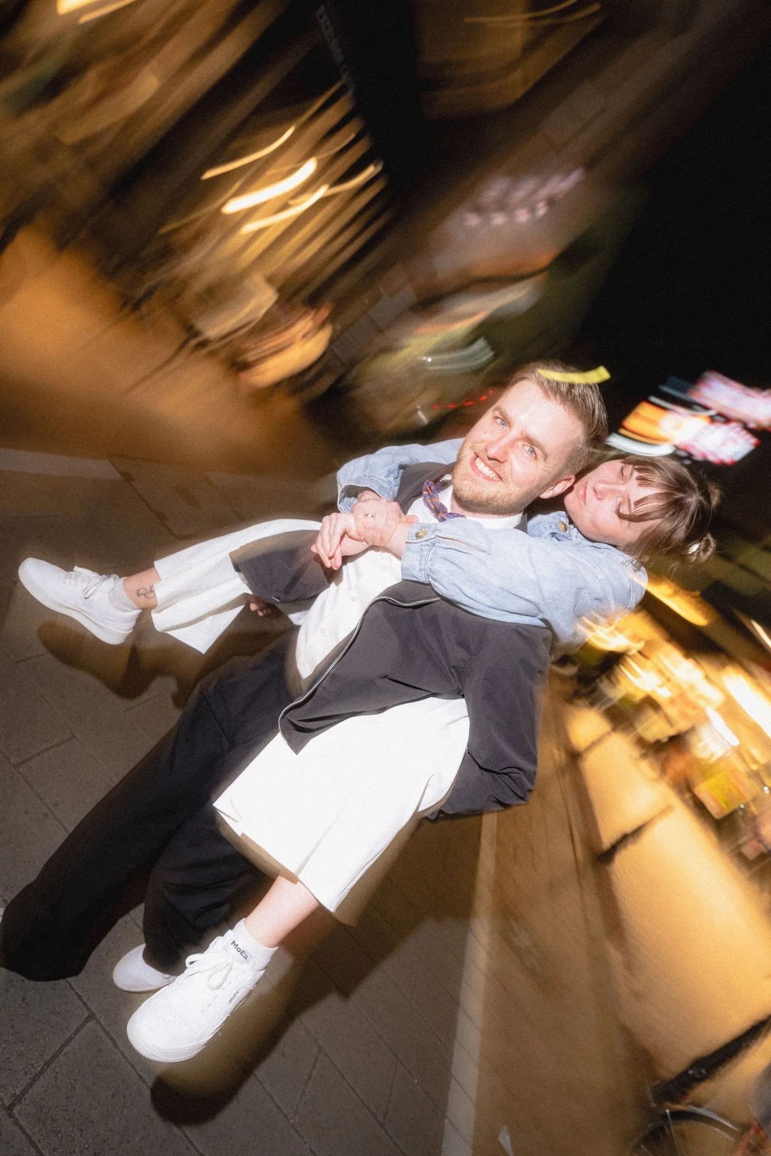 A newlywed couple smiling, one with arms around the other, in a dimly lit indoor setting with wooden decor and blurry lights in the background.