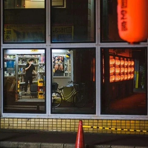A man browses shelves in a small Tokyo shop, seen through large windows reflecting red lanterns, with a single traffic cone on the sidewalk.