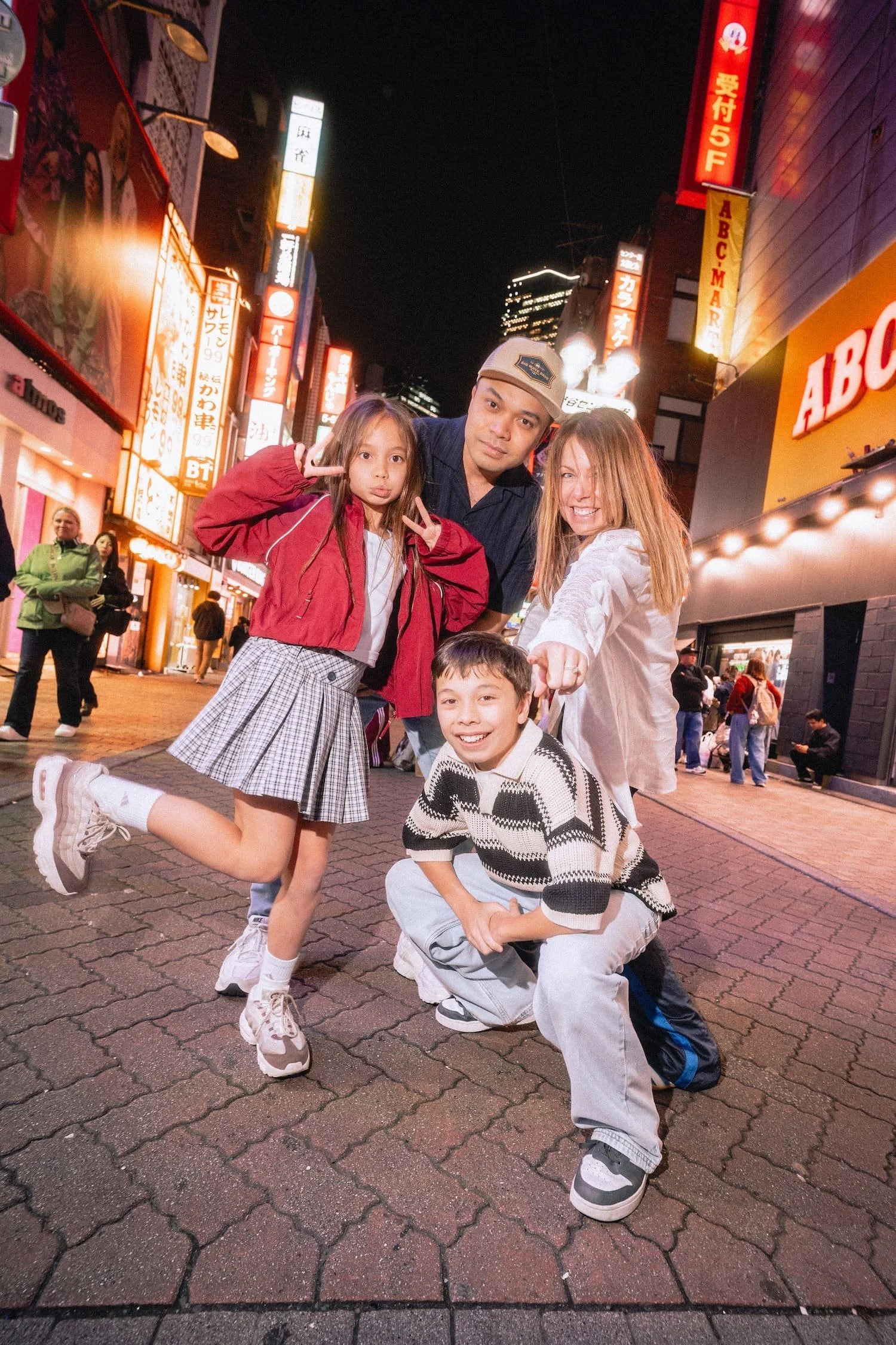 A family of four, including a woman with light brown hair, a man in a cap, a girl with long hair making a peace sign, and a boy with short hair crouching, pose for a photo at night on a busy Shibuya, Tokyo street with neon signs in the background.