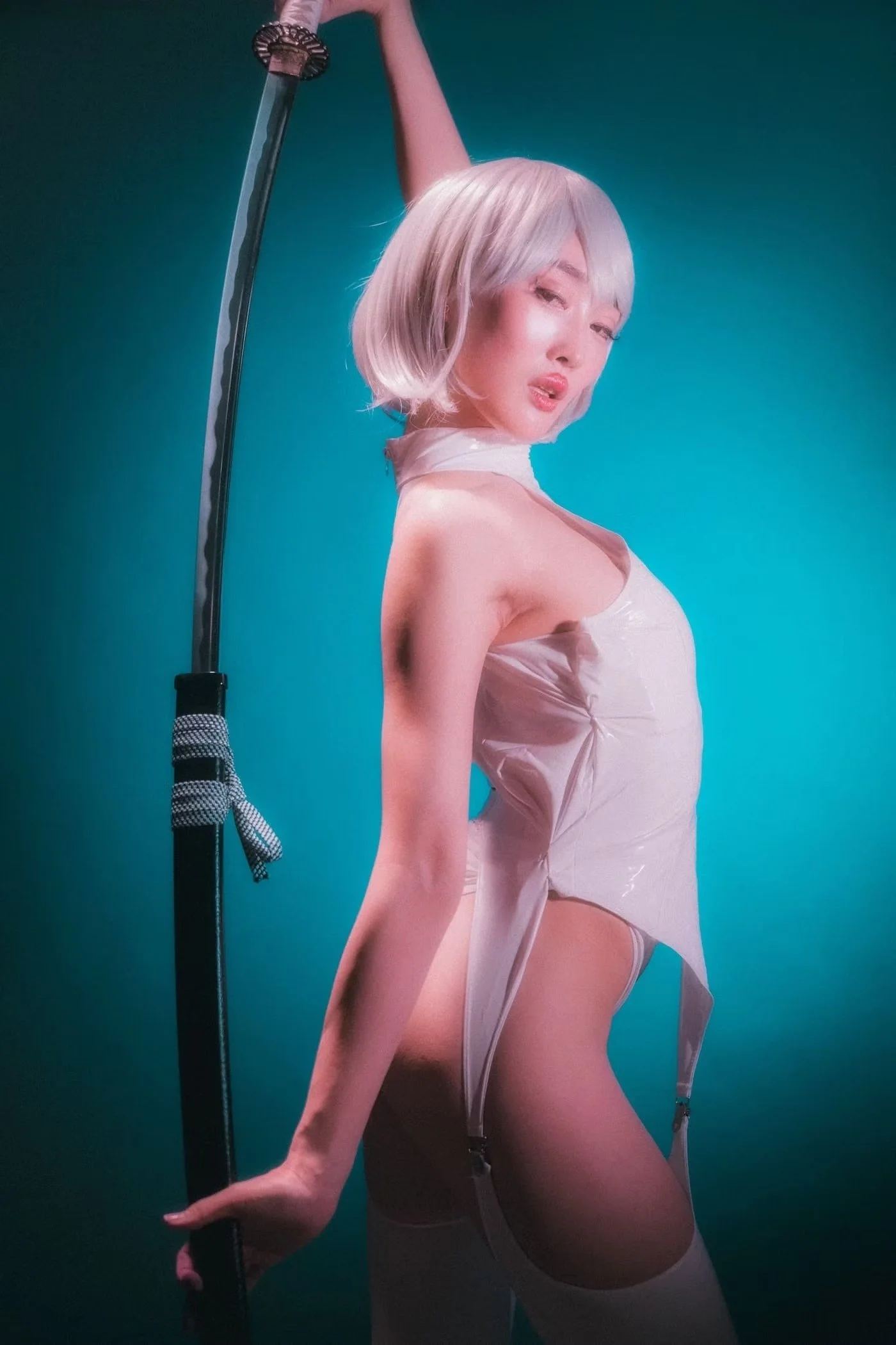 A woman with platinum blonde hair dressed in a sheer, shiny white outfit, holding a sword with a checkered cloth tied to its hilt, against a teal background.