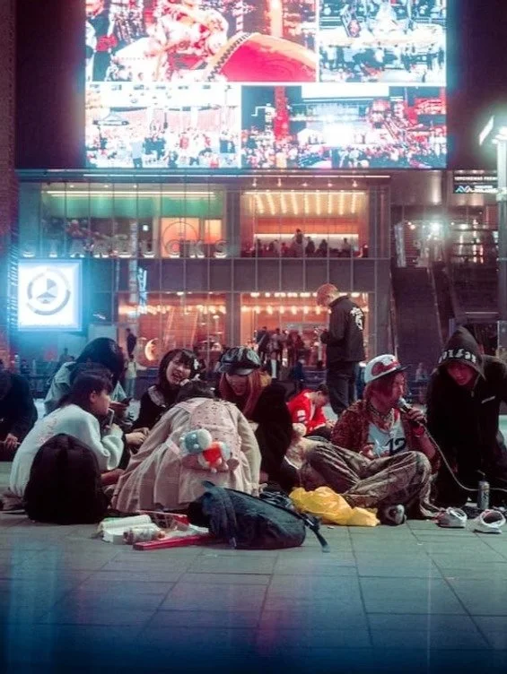 Group of people sitting on the ground in a busy urban area at night, with a large screen displaying multiple images above them and city lights in the background.