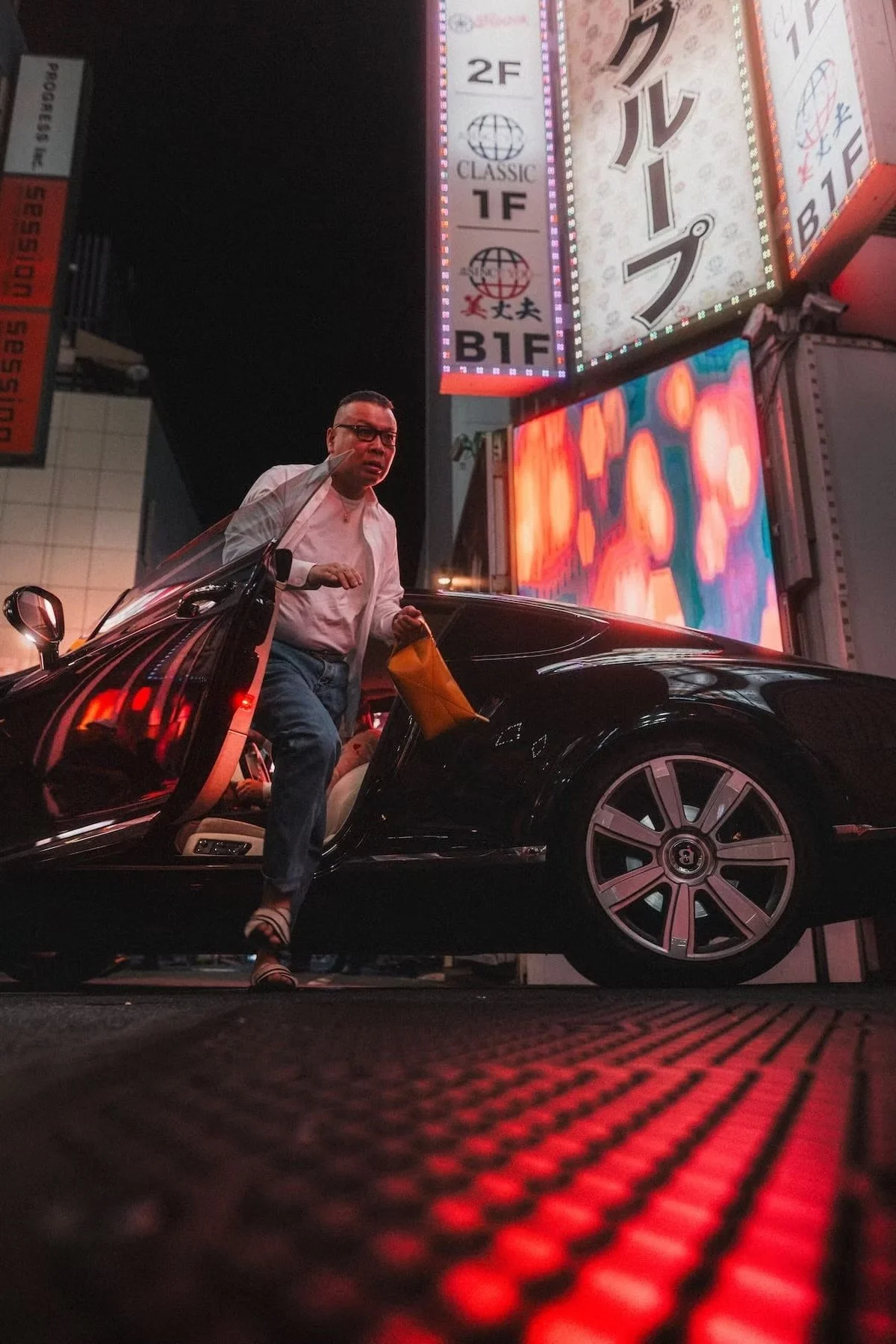 A man with glasses and casual clothing is stepping out of a black luxury sports car at night in a city with bright, colorful neon signs and billboards in the background. Kabukicho night scenes by Deniz Demir