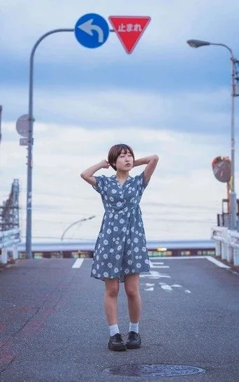 A girl in a blue dress with her arms raised, standing on a bridge in Osaka, with the city behind her.