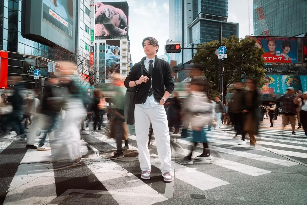 A young man standing on a busy city crosswalk among many blurred pedestrians, with tall buildings and billboards in the background.