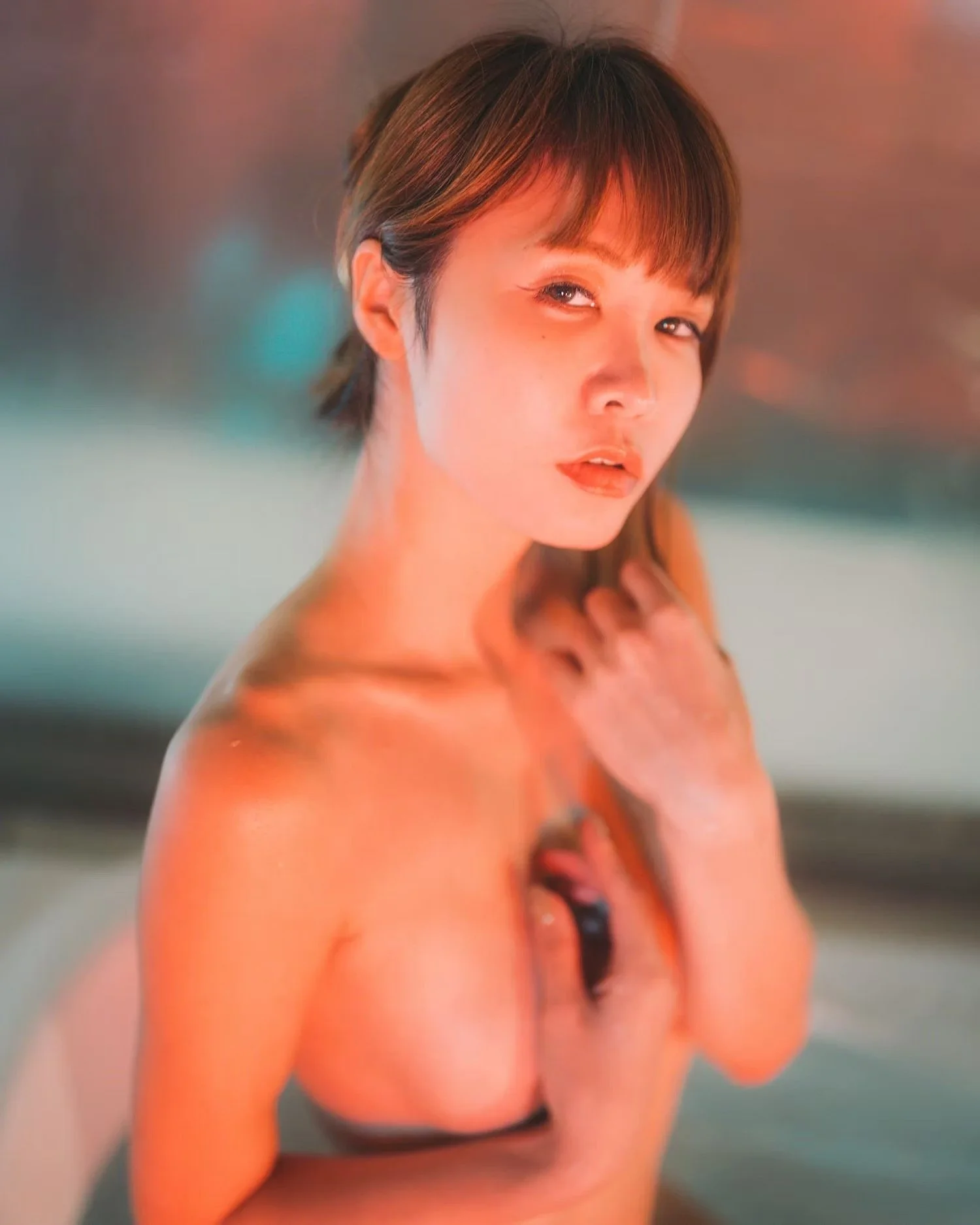 A woman with short brown hair and makeup, wearing black lingerie, posing in front of a blurry colorful background.
