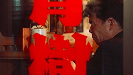 A man with dark hair and a black shirt sitting at a table in a restaurant or cafe, with bright red Chinese characters overlaid on the image.