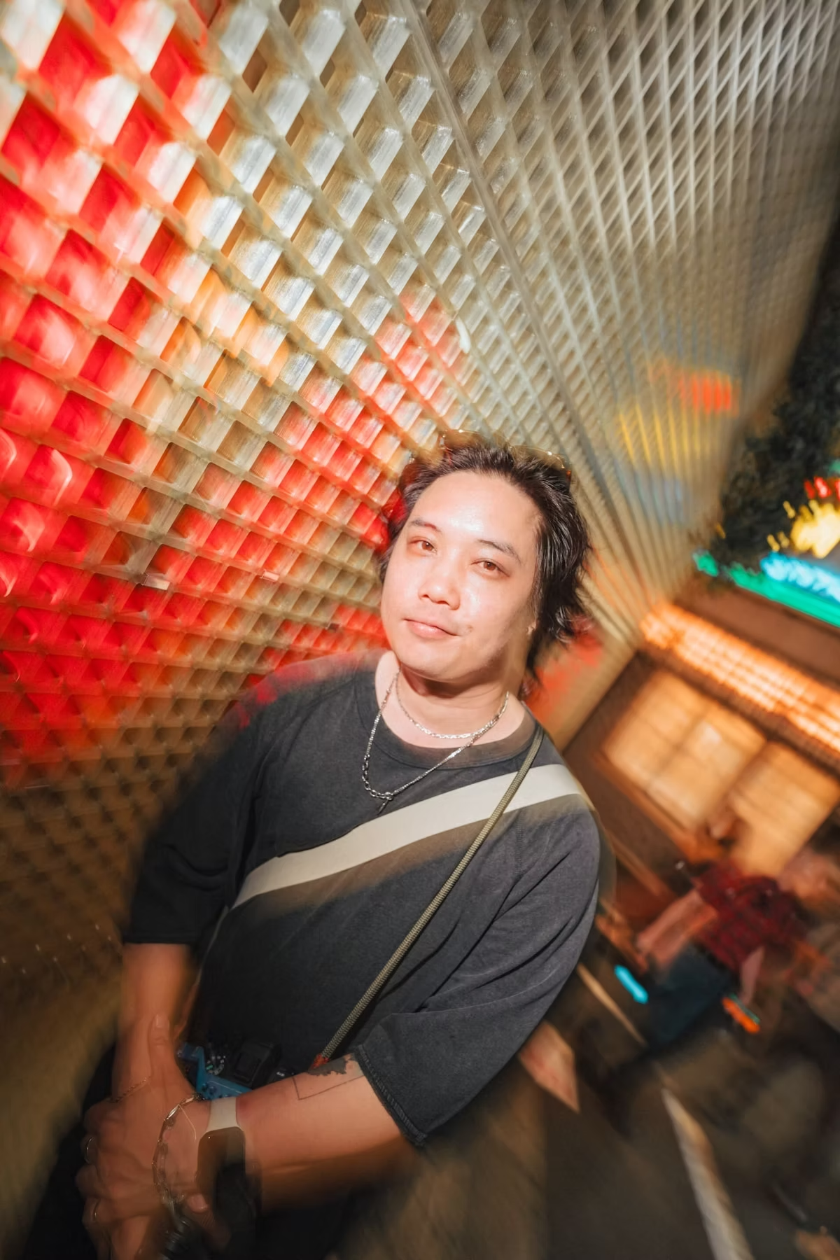 A man with a stylish look stands in front of a unique patterned background in a dimly lit urban space. The motion blur effect and the warm lighting add energy to the portrait, highlighting the vibrant city atmosphere.