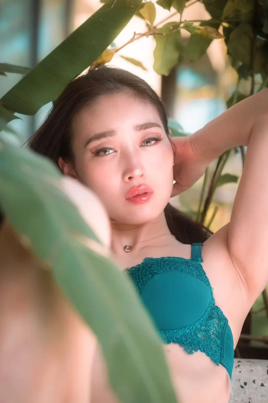 A Japanese woman with dark hair and makeup wearing a teal lace bra, posing among green leaves with her arms raised behind her head, looking into the camera.