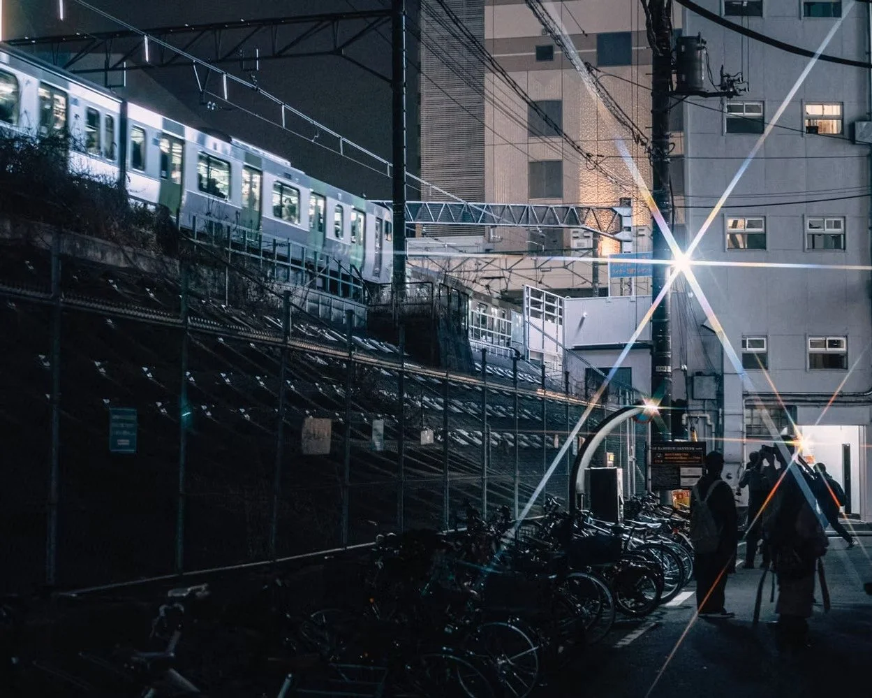 Nighttime scene of a city with a train on an elevated track, street signs, bicycles parked, and people standing in a parking area with building in the background, illuminated by bright streetlights.