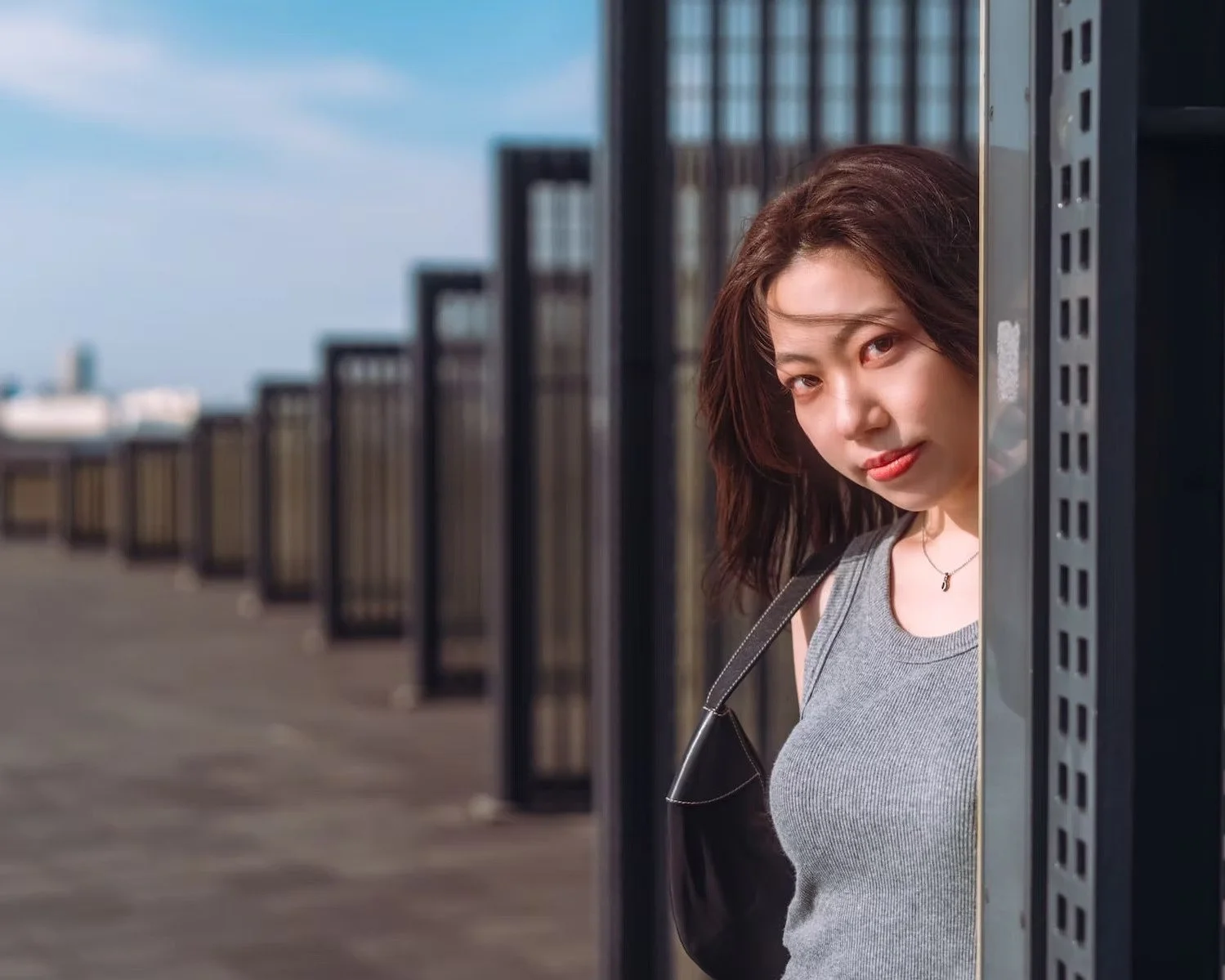 A woman with shoulder-length brown hair peeks out from behind a black metal fence on a rooftop with a city skyline in the background.