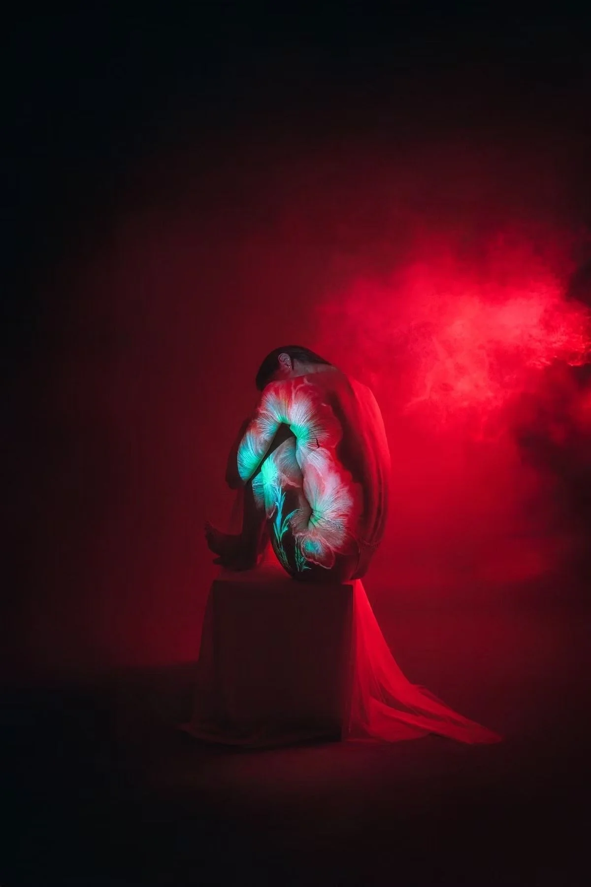 A person is sitting on a platform with their back to the camera, surrounded by red and teal smoke effects, wearing a flowing white dress, illuminated by red light in a dark setting. Studio portrait photography by Tokyo photographer Deniz Demir.
