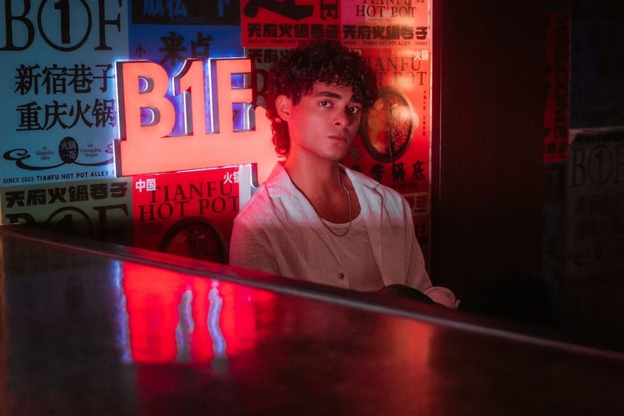 A man in a white shirt and leather jacket sits against a wall in a dark Tokyo alley, surrounded by vibrant neon signs. Neon Portraits by Tokyo Photographer Deniz Demir Shinjuku