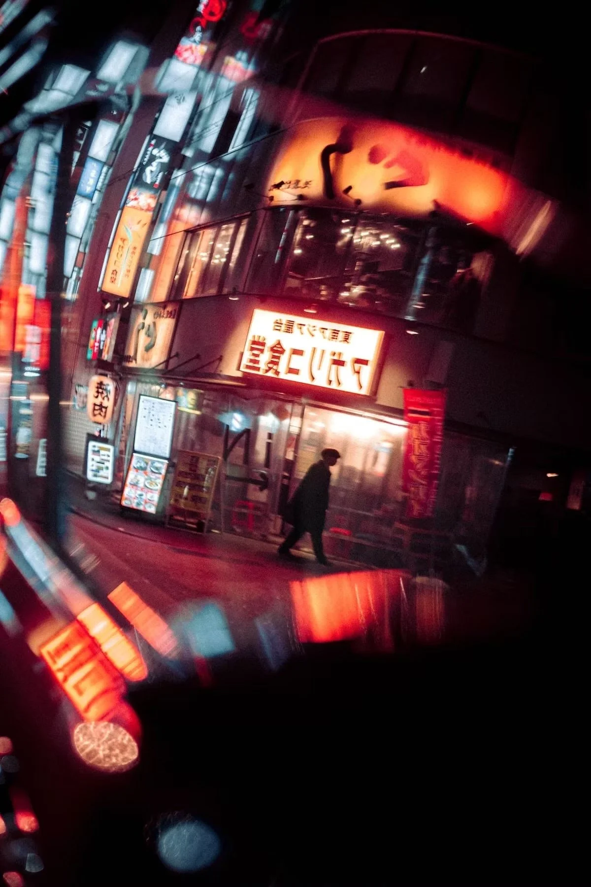 Nighttime street scene with signs in Japanese, illuminated restaurant front, and a person walking past. Reflections and blurred lights create a vibrant urban atmosphere.