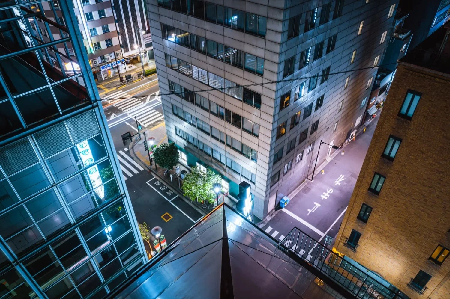 Nighttime view looking down at an urban intersection surrounded by modern buildings with illuminated windows, streetlights, and crosswalks.