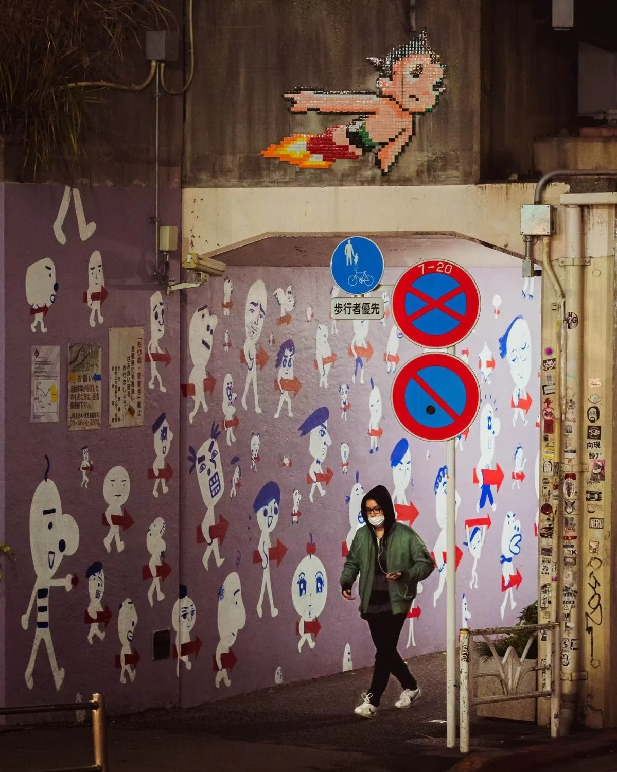 A wall decorated with Astroboy and anime characters of various facial expressions and outfits. Above, a pixel art of a flying character with a rocket, resembling a cartoon or video game sprite. A woman wearing a face mask and glasses walks in front o